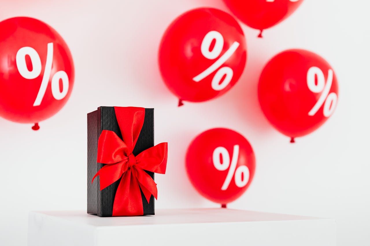 home-img Stylish black gift box with red ribbon surrounded by red percentage balloons on a white background.